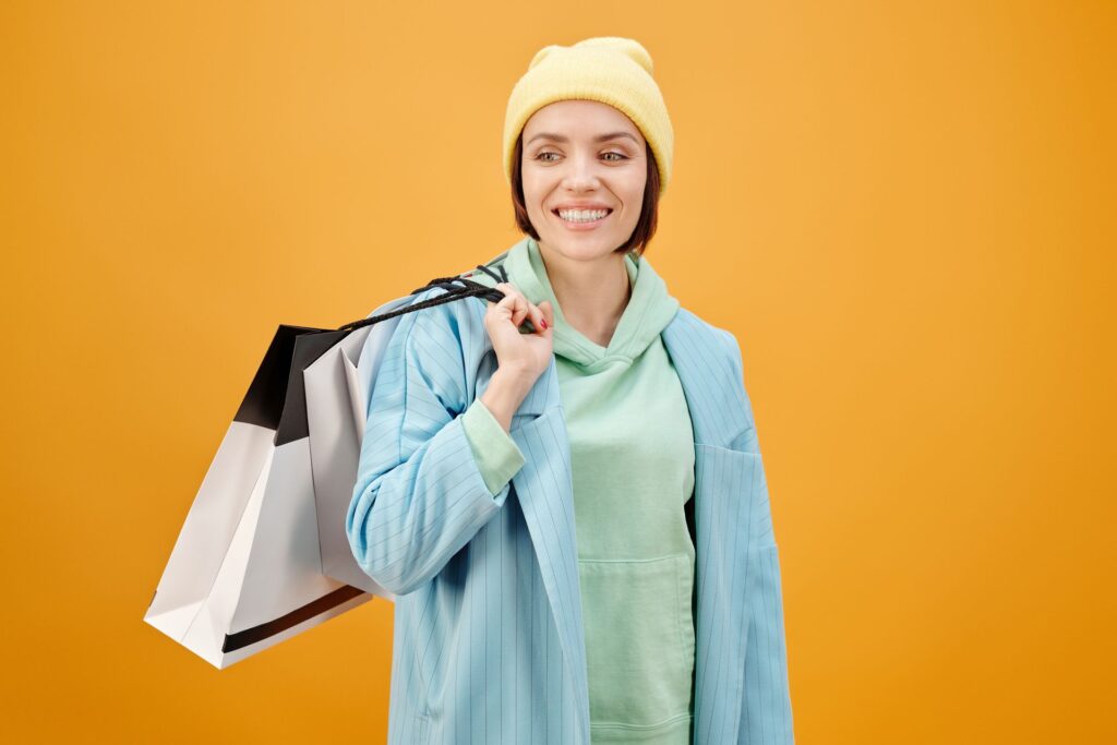 Smiling shopper wearing a yellow beanie and blue jacket, carrying shopping bags against a bright orange background.