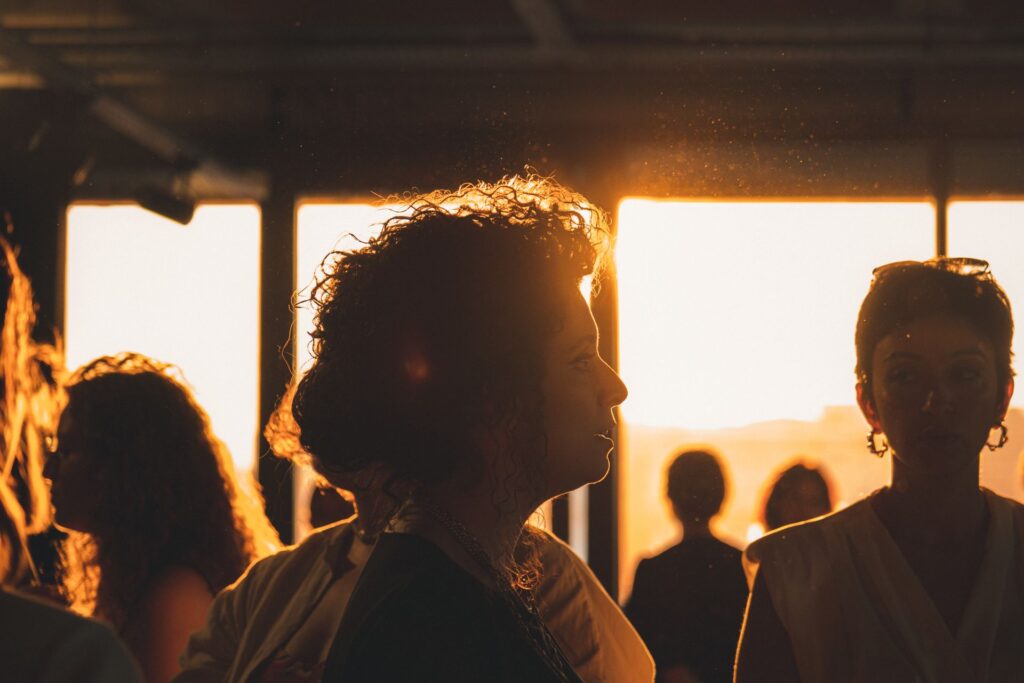People in conversation at a networking event, silhouetted against warm evening light streaming through large window, representing the connections and conversations involved in securing startup investment.